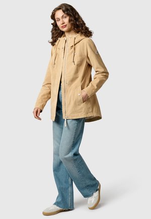 Kvinde står med den ene hånd i lommen iført tanfarvet hættetrøje, lyseblå jeans og hvide sneakers mod en ensfarvet baggrund.