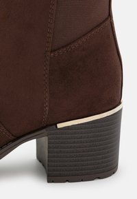 Bottine en daim marron avec une finition lisse, un bout arrondi, un talon bloc texturé avec un accent métallique, et un panneau latéral extensible.