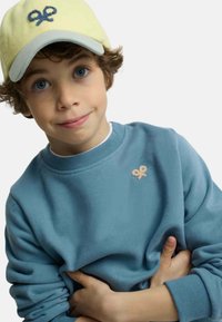 Niño pequeño con ojos azules, lleva una gorra amarilla y una sudadera azul con un pequeño logo bordado en el pecho, con los brazos cruzados delante.