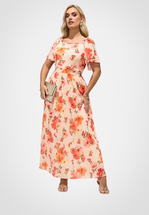 Kvinde iført en lang, ferskenfarvet blomsterkjole med flagrende ærmer, der holder en beige clutch og bærer guldfarvede sandaler med stropper samt armbånd.