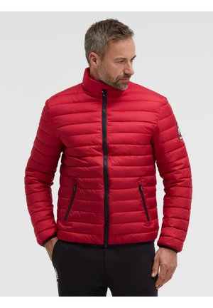 Man wearing a zipped red puffer jacket with black trim, hands in pockets, looking to the side against a plain light background.