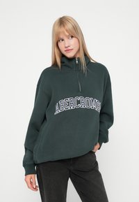 Πράσινη ζακέτα με φερμουάρ από μαλακό ύφασμα, με ψηλό γιακά και το λογότυπο "ABERCROMBIE" ραμμένο σε λευκό μπροστά, συνδυασμένη με σκούρα τζιν.