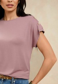 Īsām piedurknēm, gaiši mauve krāsas tops, izgatavots no mīksta auduma, ar dekoratīvām pogām uz pleca. Saskaņots ar ziliem džinsa biksēm un jostu.