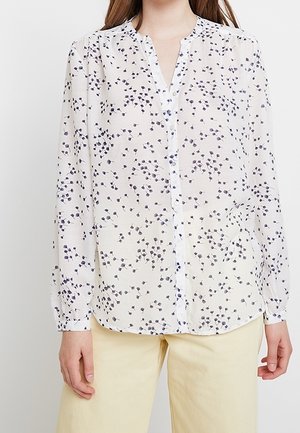 Kvinde iført en hvid langærmet skjorte med små sorte bladmønstre og lyse beige bukser, stående mod en ensfarvet baggrund.