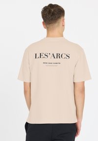 Beige kortærmet t-shirt med sort tekst på ryggen: "LES'ARCS" og "NEW BAD HABITS," med rund hals og afslappet pasform.