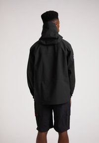 Sort vandtæt jakke med hætte, der har en afslappet pasform og subtile sømdetaljer, parret med sorte cargo shorts, alt sammen mod en ensfarvet baggrund.