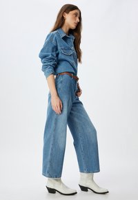 Camisa de mezclilla azul clara con cierre de botones, combinada con jeans azules holgados y un cinturón marrón, complementada con botas de tobillo blancas de estilo western.