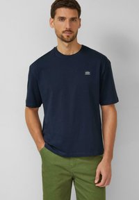 Marineblauw katoenen T-shirt met korte mouwen, ronde hals en een klein merklogo op de borst. Gecombineerd met een groene broek.