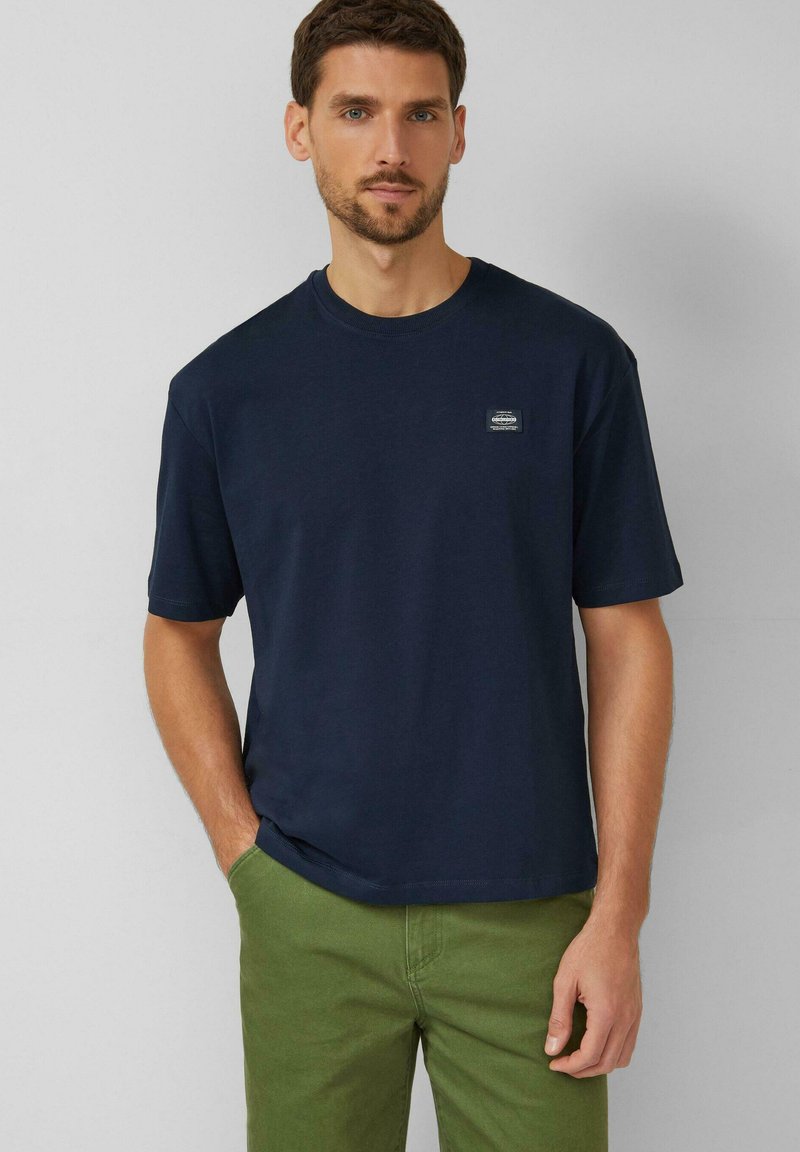 Marineblauw katoenen T-shirt met korte mouwen, ronde hals en een klein merklogo op de borst. Gecombineerd met een groene broek.
