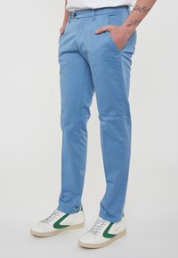 Pantaloni blu chiaro da uomo con due tasche laterali, chiusura con bottone e vestibilità dritta. Abbinati a sneakers bianche e verdi.