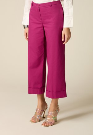 Donna che indossa pantaloni a vita alta e gambe larghe di colore magenta abbinati a sandali con tacco multicolore con cinturini e una camicia bianca a maniche lunghe.