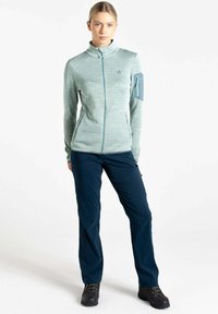 Helles grünes, langärmliges Zip-Jacket mit hohem Kragen und Arm-Tasche, kombiniert mit dunkelblauen Hosen und schwarzen Outdoor-Schuhen.