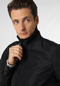 Jachetă neagră din softshell cu guler înalt, închidere cu fermoar, textură netedă și detalii de cusătură subtile. Modelul ține fermoarul cu o mână.