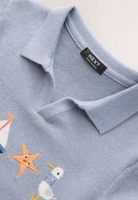 Lyseblå strikket polo med broderede motiver af en sejlbåd, en søstjerne og en måge. Mærket viser "NEXT, lavet i Indien."
