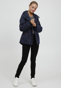 Marineblauwe waterdichte jas met een capuchon, voorzien van twee zijzakken, metalen details en een ritssluiting. Gecombineerd met zwarte slim-fit broek en witte sneakers.