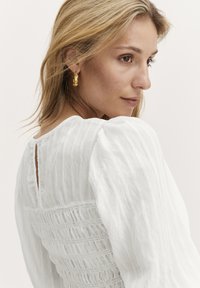 Vit texturerad blus med puffärmar och smockad rygg. Guldörhängen i ringform framhäver halslinjen. Elegant och enkelt design.