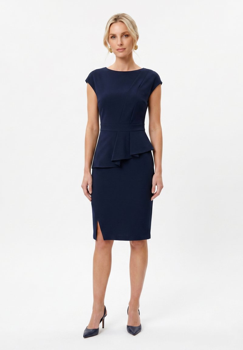 Femme portant une robe bleu marine cintrée avec manches courtes, détail péplum et fente à l'avant, associée à des talons hauts bleu marine et des boucles d’oreilles dorées.