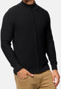 Sudadera de punto negra con relieve, con cierre de cremallera y cuello alto. Su diseño ajustado resalta la parte superior del cuerpo, con una textura suave.