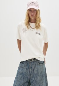 Kvindelig iført en hvid T-shirt med blomster- og teksttryk, en lys pink "Positano Italy" kasket, lag på lag halskæder og løse blå jeans med et naglet bælte.