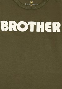Žalias medvilninis marškinėlis su didelėmis baltomis blokinėmis raidėmis, kurios rašo "BROTHER" ant priekinės dalies. Apvalus iškirpimas ir trumpomis rankovėmis.