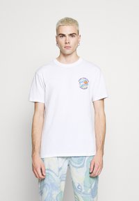 Hvid bomulds t-shirt med rund halsudskæring og korte ærmer, med et grafisk tryk i blå og sort på venstre brystområde.