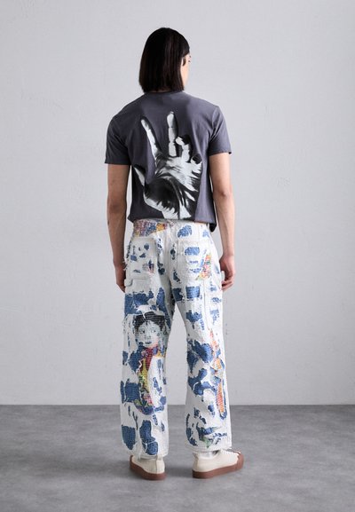 Gray t-shirt featuring a large black hand graphic on the back; white, patterned cargo pants with colorful imagery; cream shoes.