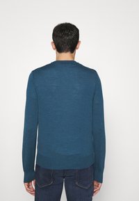 Man standing facing away wearing a teal long-sleeve sweater and dark blue jeans against a plain white background.