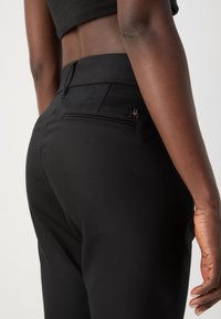 Gros plan d'une personne portant un pantalon noir ajusté et un crop top noir, montrant les détails de dos et de côté avec un petit logo métallique sur la poche.