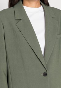 Veste blazer vert surdimensionnée en tissu texturé avec un col cranté et un bouton noir unique. Portée sur un t-shirt blanc à col rond.