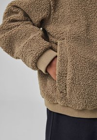 Brun, fluffy fleece pullover med en struktureret overflade, ribbede manchetter og sidelommer. Den nederste kant er beige, hvilket komplimenterer hovedfarven.