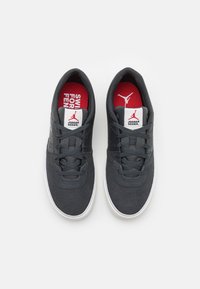 Mörkgrå sneakers gjorda av strukturerat material med rund tå, vit sula och svarta snören. Röda innersulor har logotyp och text.