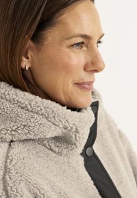 Fleece jack in lichtgrijs met een gestructureerde afwerking, voorzien van een hoge kraag en zwarte knoopsluiting, gedragen met kleine zilveren oorbellen.