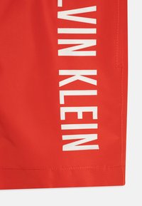 Røde svømmetight laget av lett materiale med et hvitt logotrykk langs siden, med rett snitt og sømdetaljer.