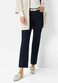 Jeans de mezclilla azul oscuro de pierna recta, usados con un suéter claro y un cinturón beige. El calzado presenta mocasines de color claro con acentos plateados.