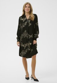 Blondinka u crnoj haljini s dugim rukavima ukrašenoj bež cvjetnim uzorcima i crnim dugim sandalama s niskom petom, stoji na bijeloj pozadini.