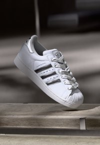 Baskets Adidas Superstar blanches avec des rayures argentées et un bout en coquillage flottant au-dessus d'une surface en pierre à l'intérieur.