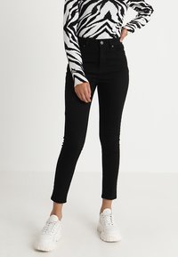 Pantalones vaqueros negros de talle alto con un corte ajustado y un ligero estiramiento. Combinados con una blusa de manga larga a rayas zebra en blanco y negro y zapatillas blancas.