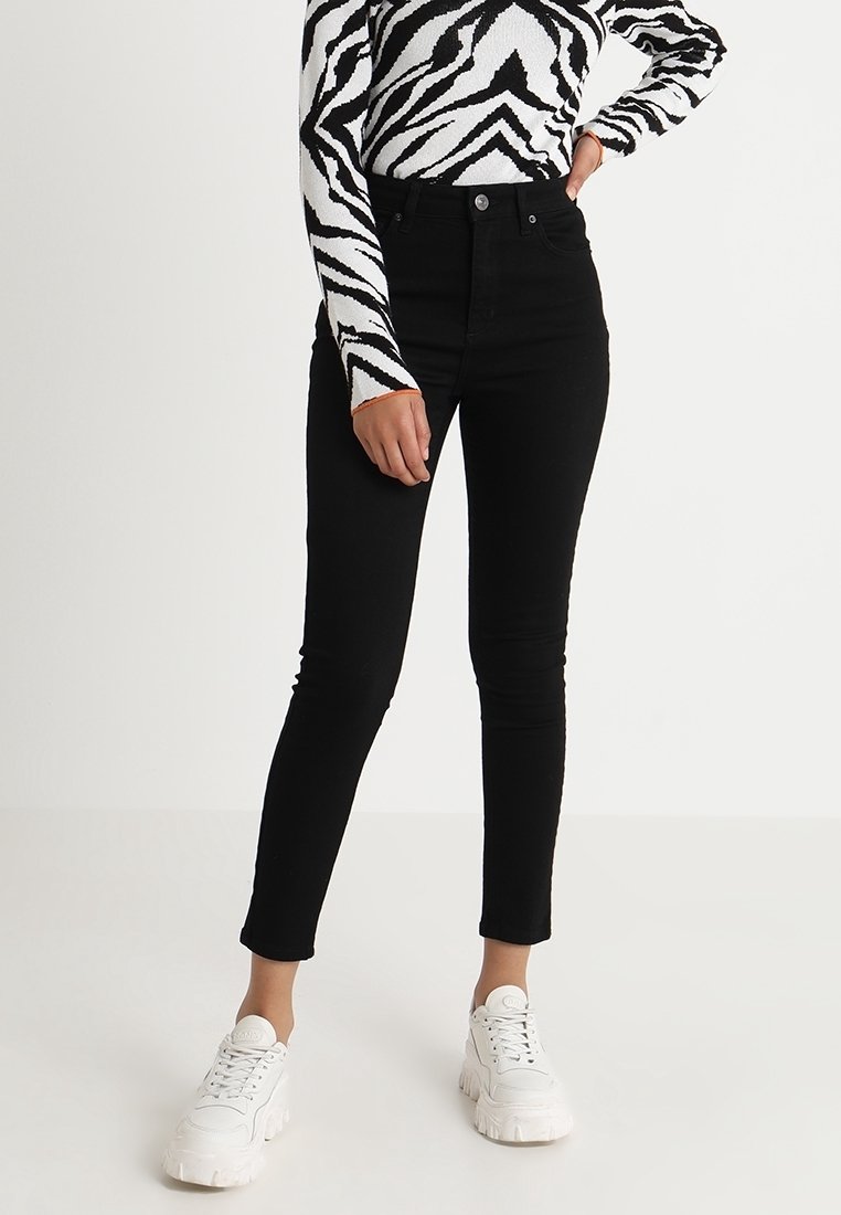 Pantalones vaqueros negros de talle alto con un corte ajustado y un ligero estiramiento. Combinados con una blusa de manga larga a rayas zebra en blanco y negro y zapatillas blancas.