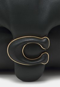 Musta nahkainen käsilaukku, jossa on veistoksellinen muotoilu ja n�äyttävä kullanvärinen pyöreä solki. Kuvioitu pinta ja sileät yksityiskohdat.