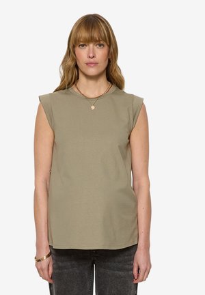 Frau mit glatten blonden Haaren und Pony, trägt ein ärmelloses beiges Oberteil, schwarze Jeans, goldene Halskette und Armband, schaut direkt in die Kamera vor weißem Hintergrund.