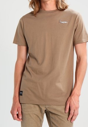 Homme portant un t-shirt marron uni avec un petit motif d'avion près de l'épaule gauche et un pantalon marron assorti.