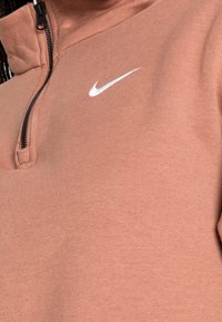 Bronsfärgad kvartzip-tröja i mjukt tyg, med en svart dragkedja och en vit Nike-logotyp på bröstet. Inga synliga mönster eller detaljer.