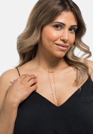 Collier en or doré avec une barre horizontale et deux pendentifs verticaux. Le modèle porte un débardeur noir, mettant en valeur la longueur et le design du collier.