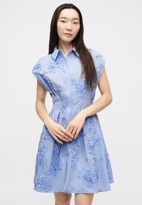 Γυναίκα που φοράει αμάνικο ανοιχτό μπλε φόρεμα με κεντήματα λουλουδιών, στέκεται μπροστά σε απλό λευκό φόντο.