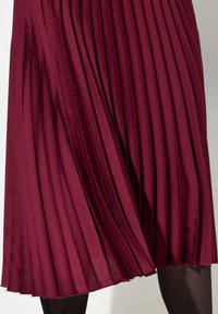 Burgundy plisserad kjol med en slät, glansig yta. Plisseringarna skapar en flödande design, fållen har en ren finish. Svarta strumpbyxor bärs under.