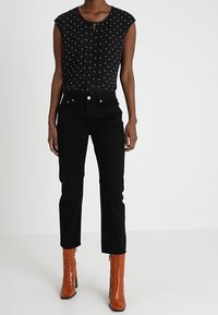 Femme portant un haut sans manches noir à pois blancs, un jean noir taille haute et des bottines brunes brillantes, se tenant devant un fond blanc.