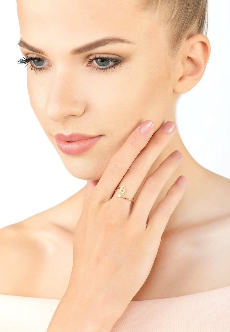Złoty pierścionek z geometrycznym wzorem w kształcie litery "B" oraz małym akcentem w postaci gwiazdki. Gładka, wypolerowana powierzchnia, noszony na dłoni z jasnoróżowym manicurem.