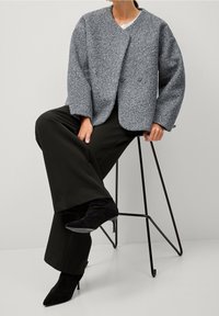 Personne portant une veste grise texturée, un pantalon noir à jambes larges et des talons noirs à bouts pointus, assise sur un tabouret minimaliste en métal noir.