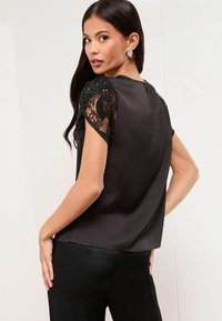 Čierna blúzka s čipkovými krátkymi rukávmi a hladkým saténovým telom. Má zaoblený lem a uzáver na zadnom výstrihu.