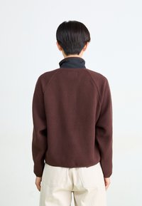 Persona con pelo corto y oscuro, vistiendo una chaqueta de forro polar marrón oscuro y pantalones de color beige claro, de pie frente a un fondo blanco.